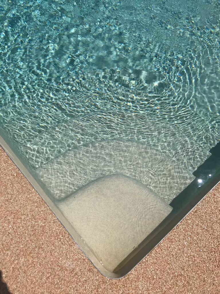 Corner of a glass-walled pool with clear, rippling blue-green water and a curved concrete edge, bordered by a pebbled pool deck.