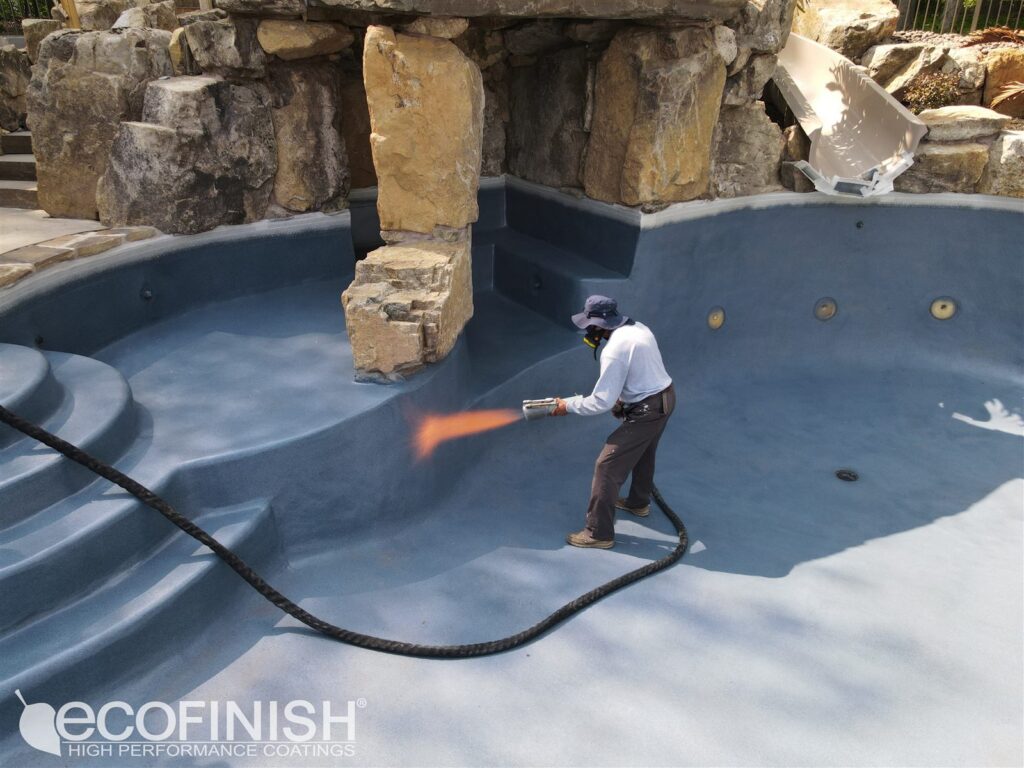 Worker wearing a hat and mask spraying orange coating inside a blue, rocky pool area using a handheld nozzle.